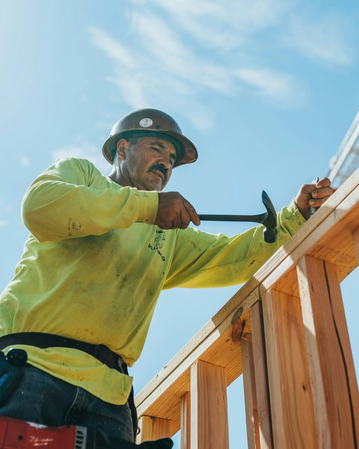 Een timmerman met helm slaat een spijker in een houten regelwerk, tegen een blauwe lucht