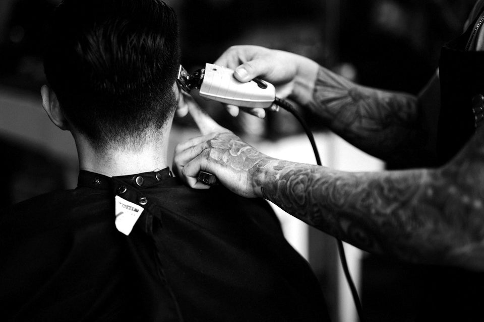 Zwart-witfoto: een barbier met getatoeëerde armen werkt de nek bij met een tondeuse, de klant van achteren gezien
