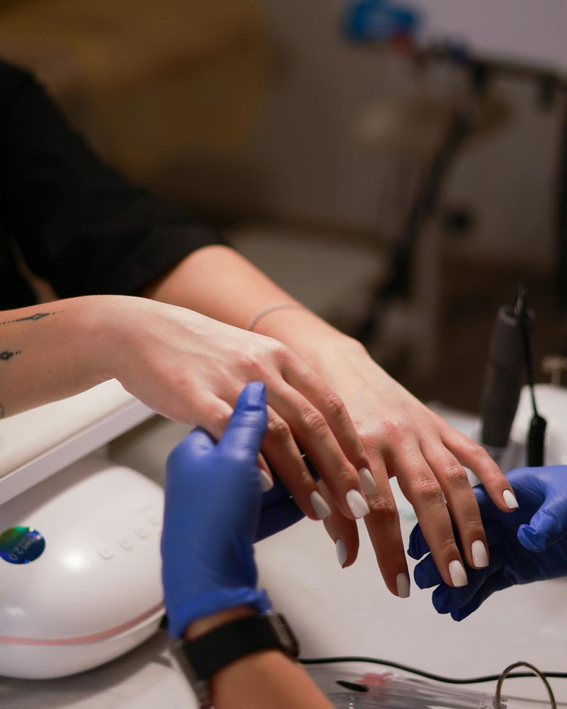 Een styliste met blauwe handschoenen werkt aan witte nagels naast een ledlamp op de werktafel