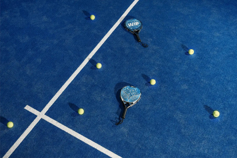 Twee padelrackets en gele ballen op een blauwe baan, van bovenaf