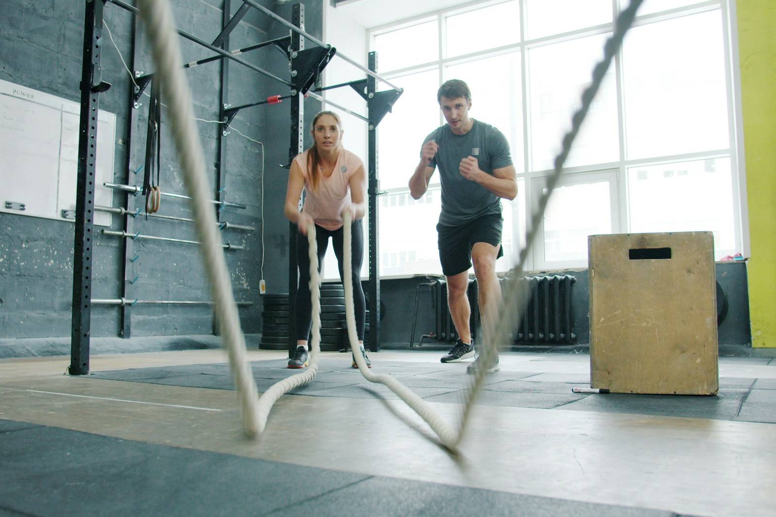 Een vrouw traint met battle ropes in een lichte sportschool met grote ramen, een trainer doet achter haar een oefening voor