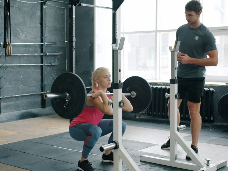 Een sporter doet een squat met een halter in het rek, de trainer staat ernaast en kijkt naar haar houding