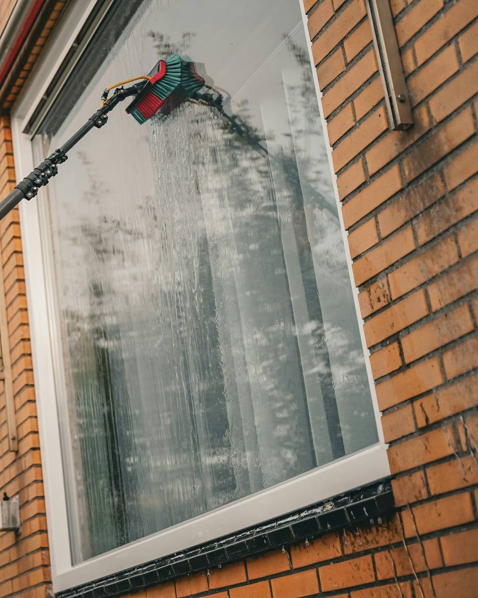 Een raam wordt met een telescoopsteel en zuiver water gewassen bij een bakstenen woning