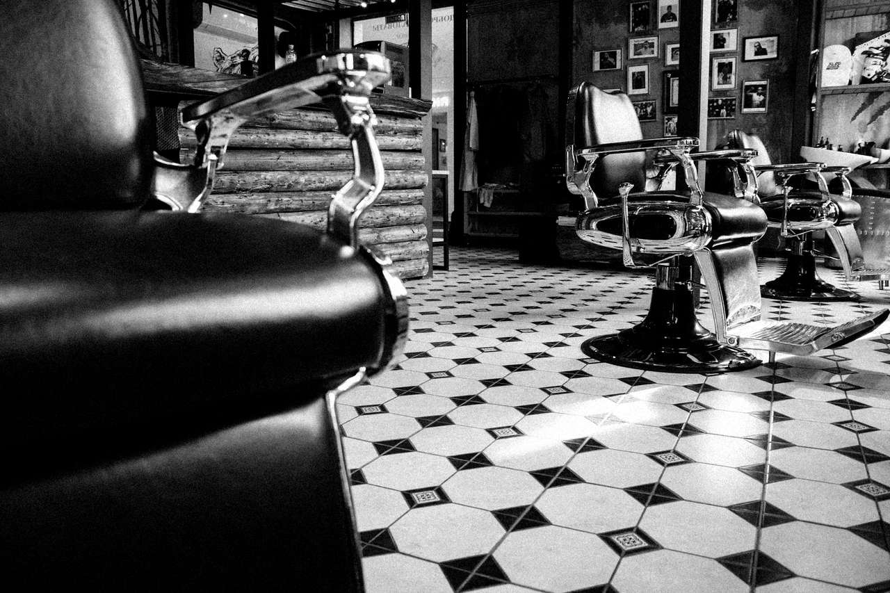 Zwart-witfoto van een lege leren kappersstoel, daarachter een rij stoelen op een tegelvloer